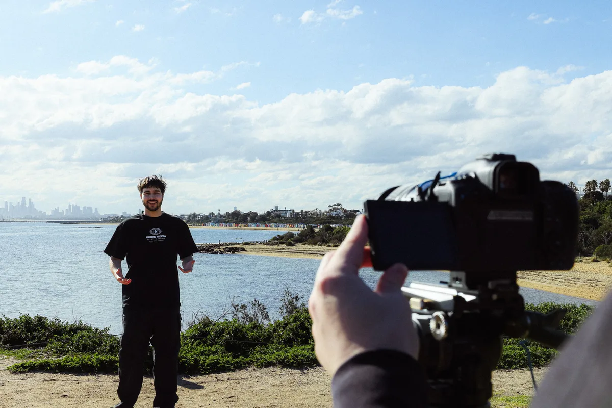Gabe presenting to camera at Brighton Beach, Melbourne skyline behind