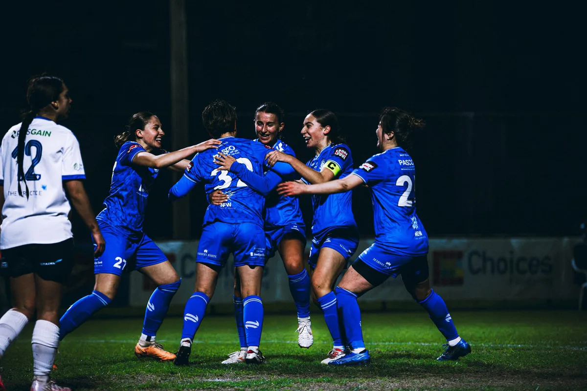 Box Hill United players in blue celebrate a goal together under lights.