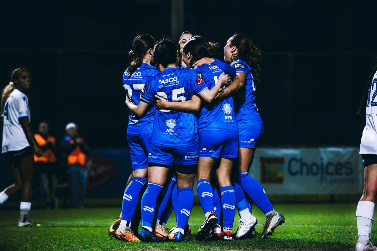 Box Hill United players in a tight huddle, celebrating a goal at night.