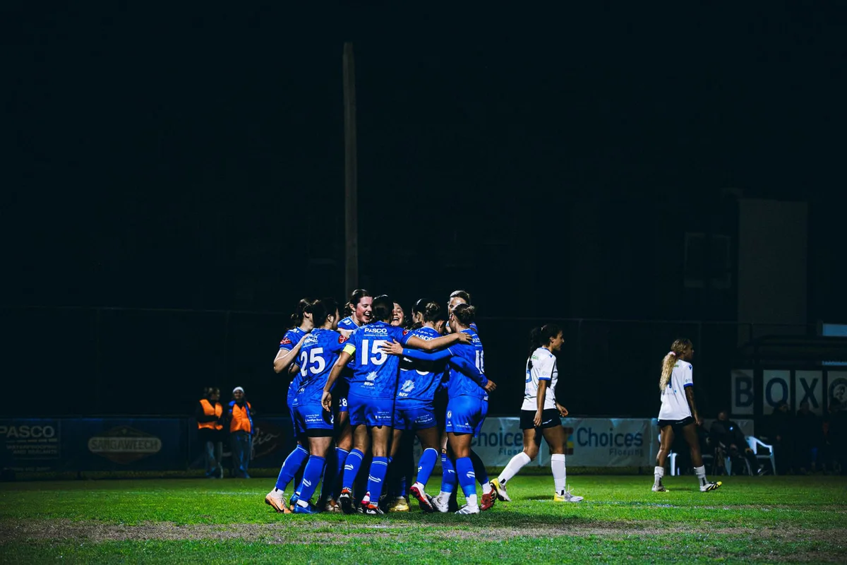 A wide night shot of Box Hill United players celebrating on the pitch.