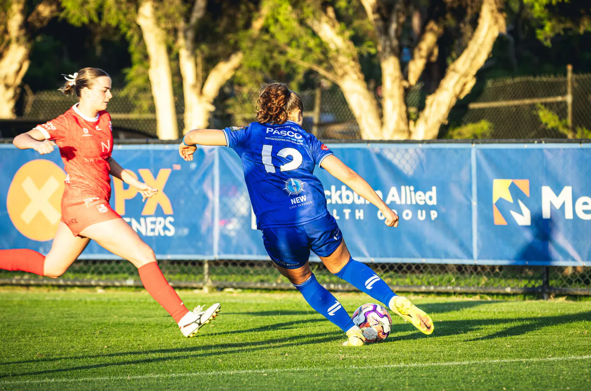 A Box Hill United player strikes the ball as a keeper in orange dives, sponsor boards behind.