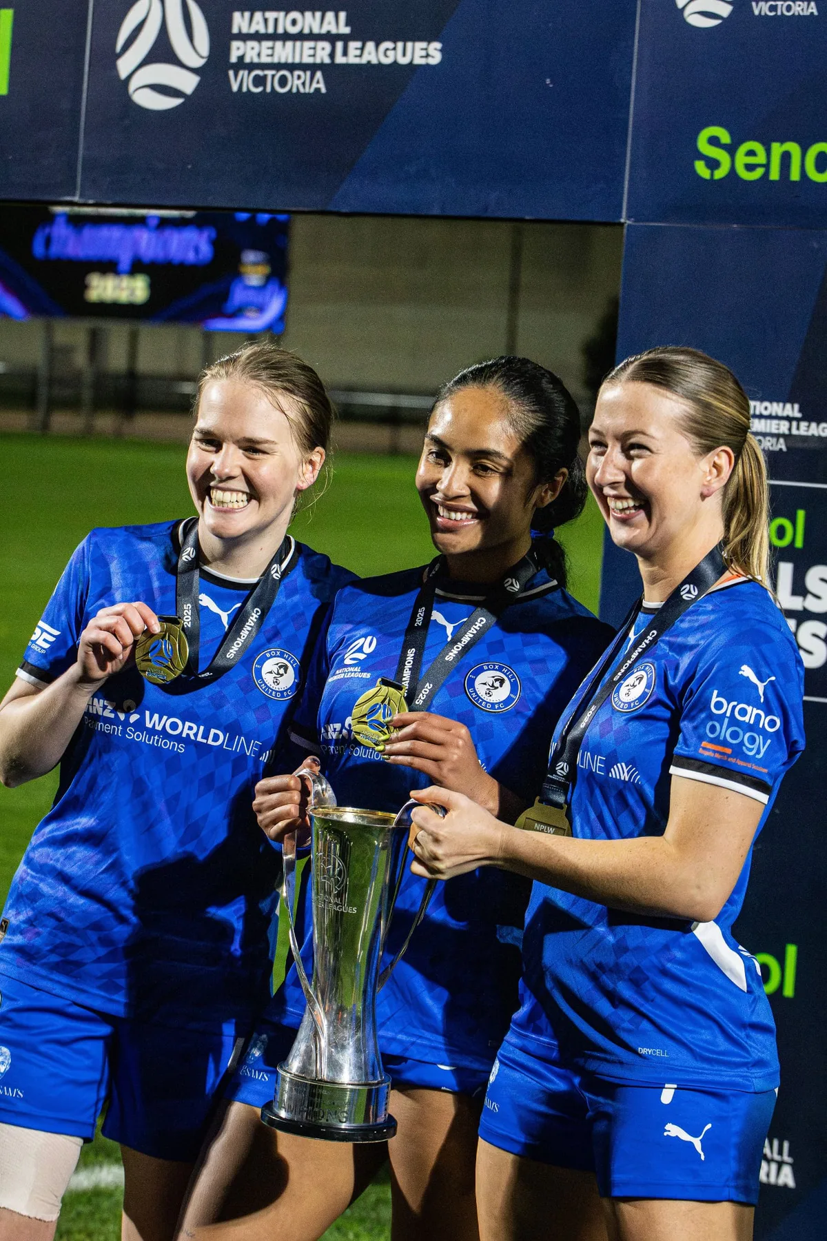 Three Box Hill United players with medals smile beside the NPL Victoria trophy in front of the presentation board.