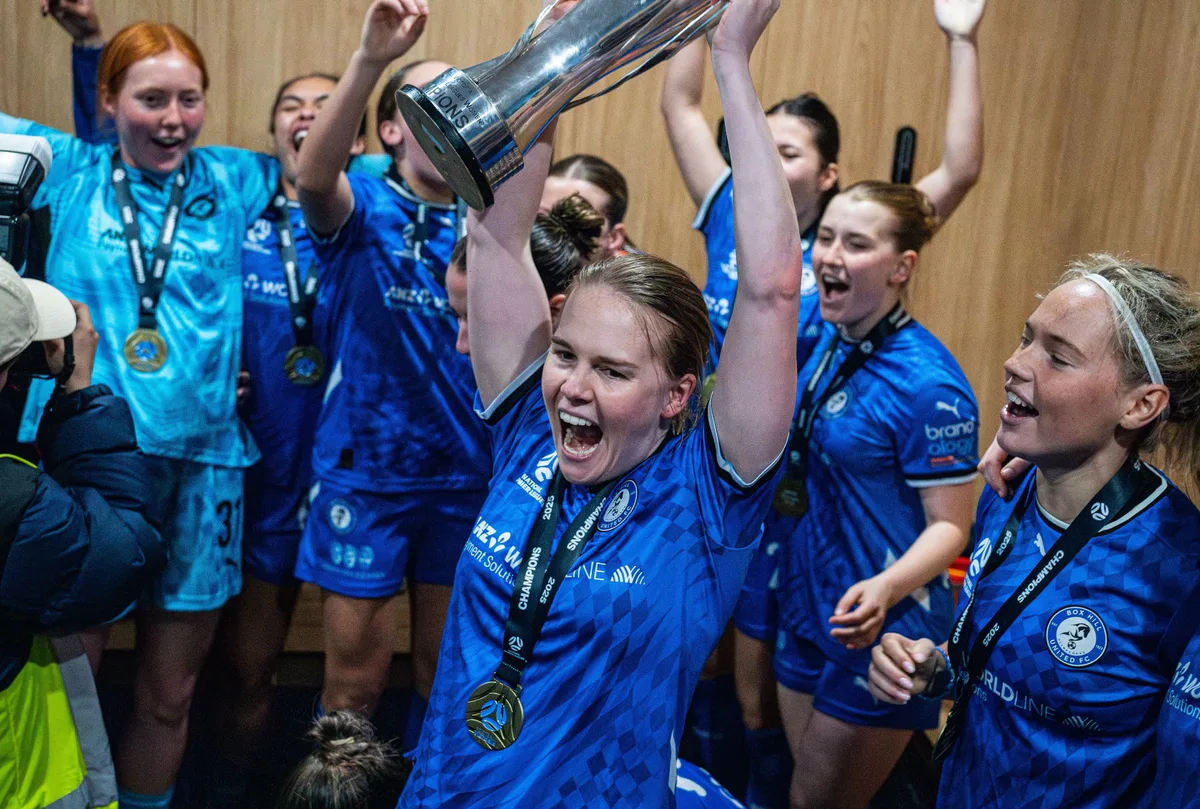 Box Hill United players celebrate in the change rooms, one raising the trophy overhead.