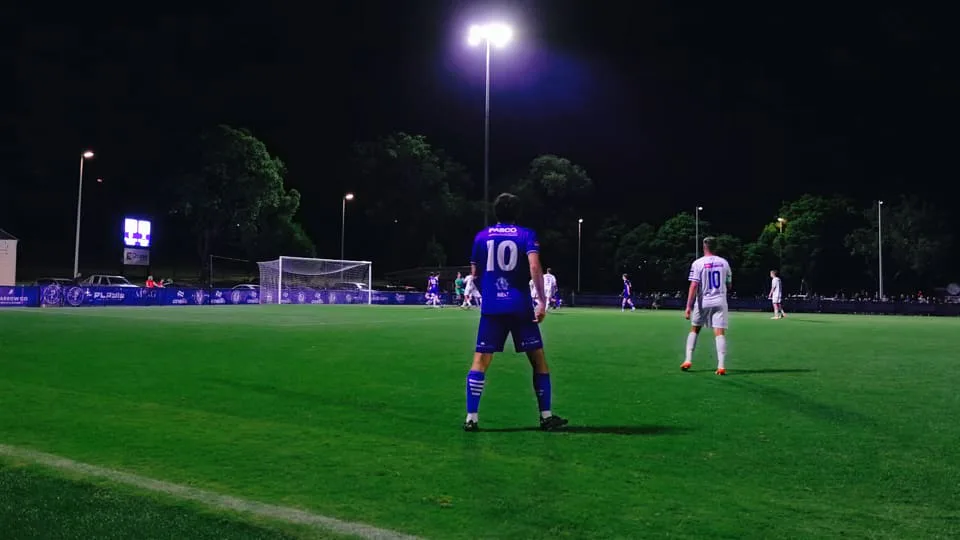 A Box Hill United player in blue standing on the floodlit pitch at night