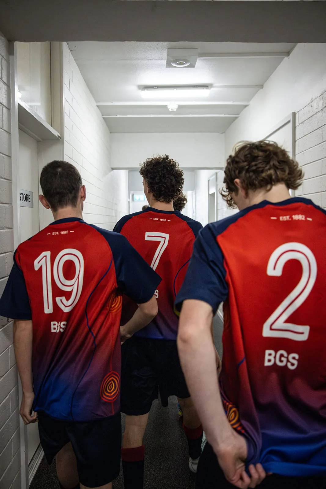 Brighton Grammar players in numbers 19, 7 and 2, seen from behind, in the corridor before a match.