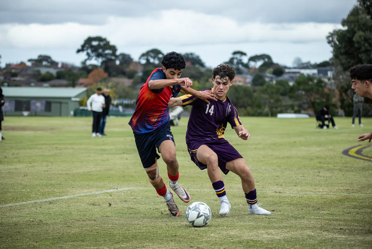 A Brighton Grammar First XI player in red and navy battles an opponent in purple for the ball.