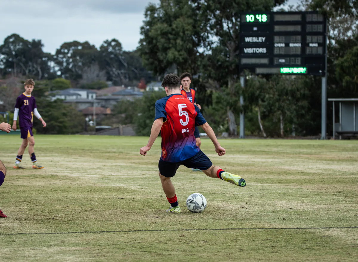 Brighton Grammar First XI players in red and navy chase the ball in a 2025 match.
