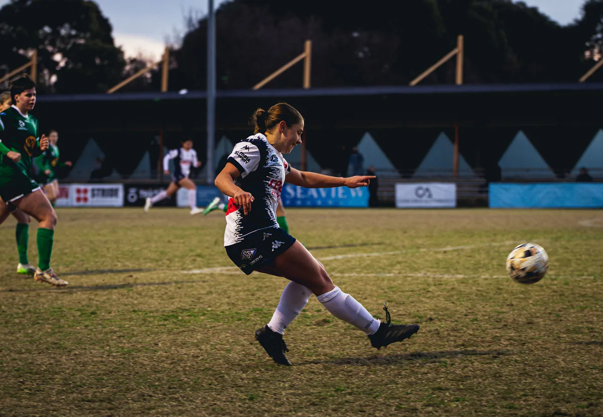 A Calder United player in white plays the ball forward under the trees.