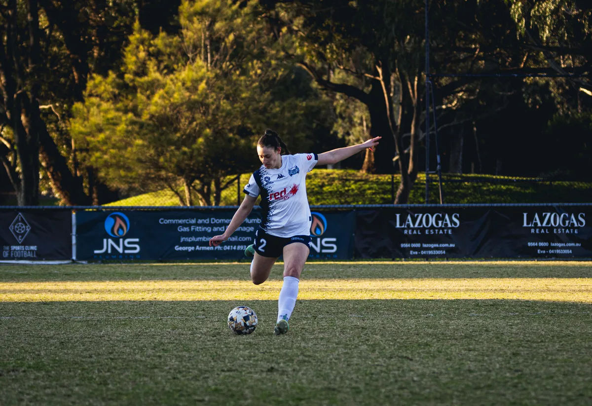 A Calder United player in white lines up a kick in late afternoon light, trees behind.