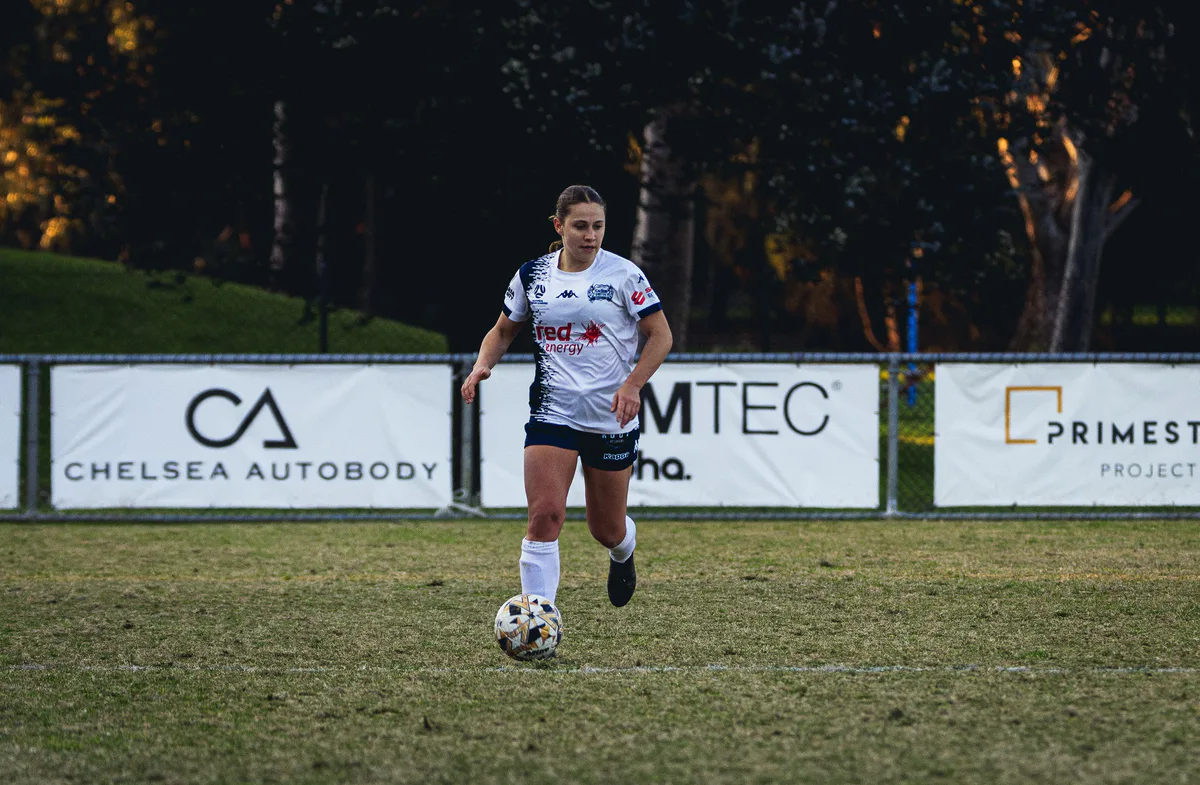 A Calder United player in white runs with the ball, sponsor boards behind.