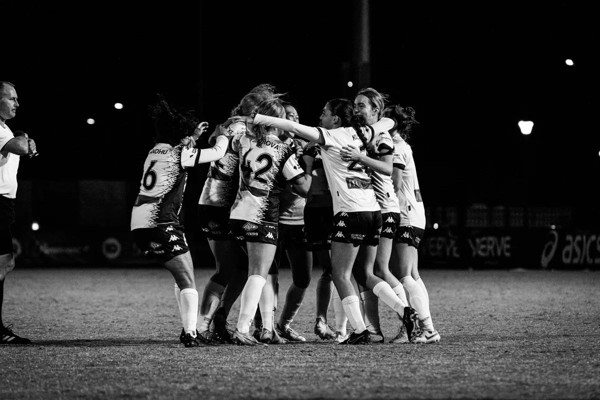 Black and white of Calder United women celebrating in a tight group on the pitch at night
