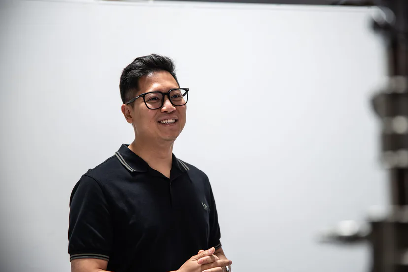 A man in glasses and a black polo smiling against a white backdrop