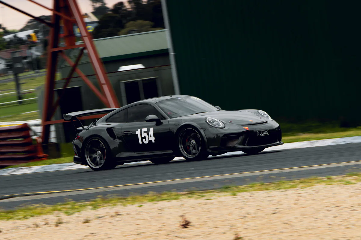 A dark grey Porsche, number 154, at speed past a trackside gantry, the kerb and grass blurred.