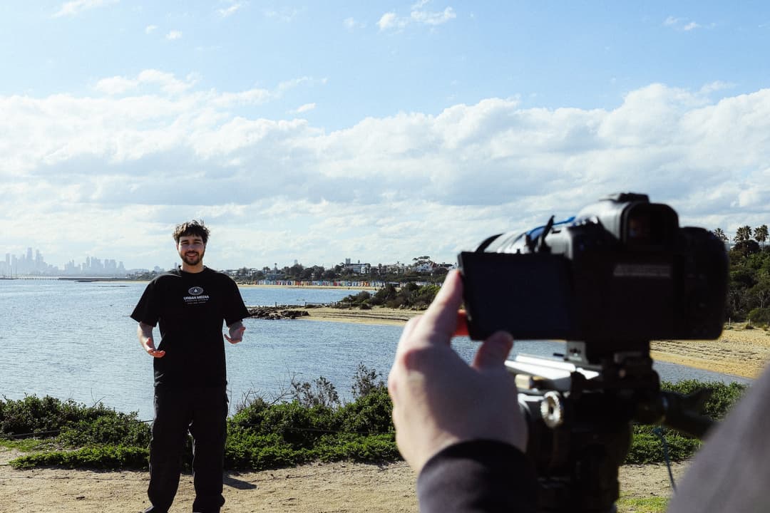 Gabe presenting to camera at Brighton Beach, Melbourne skyline behind