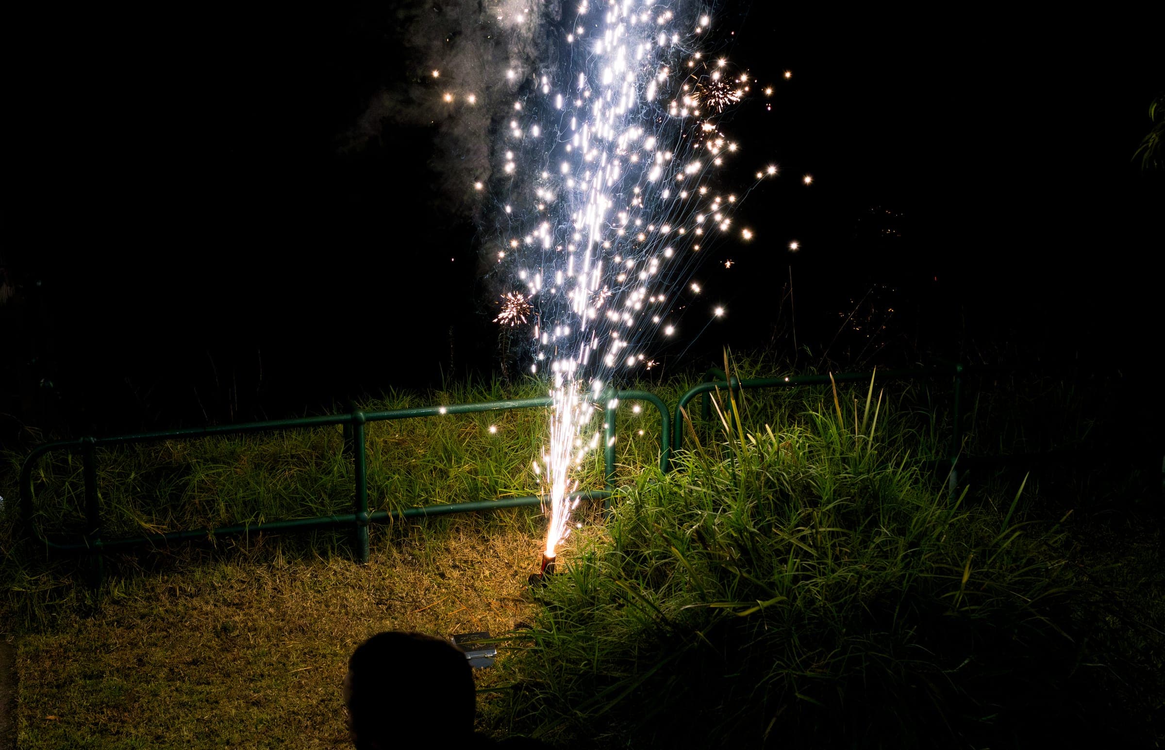 A ground fountain firing white sparks from a lawn behind a fence at night