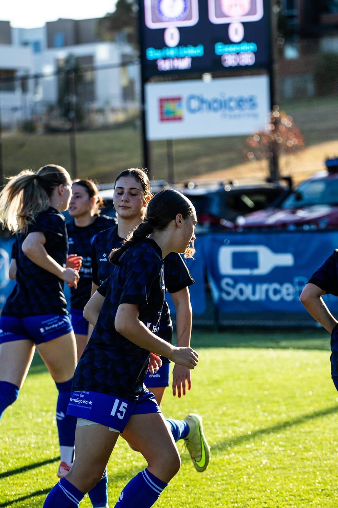 Box Hill United women warming up in training tops, the ground's scoreboard behind them