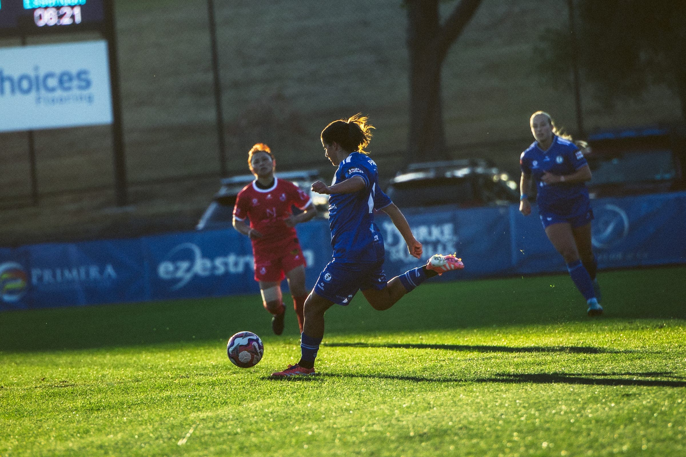 A Box Hill United player striking the ball at a floodlit match, opponent closing in