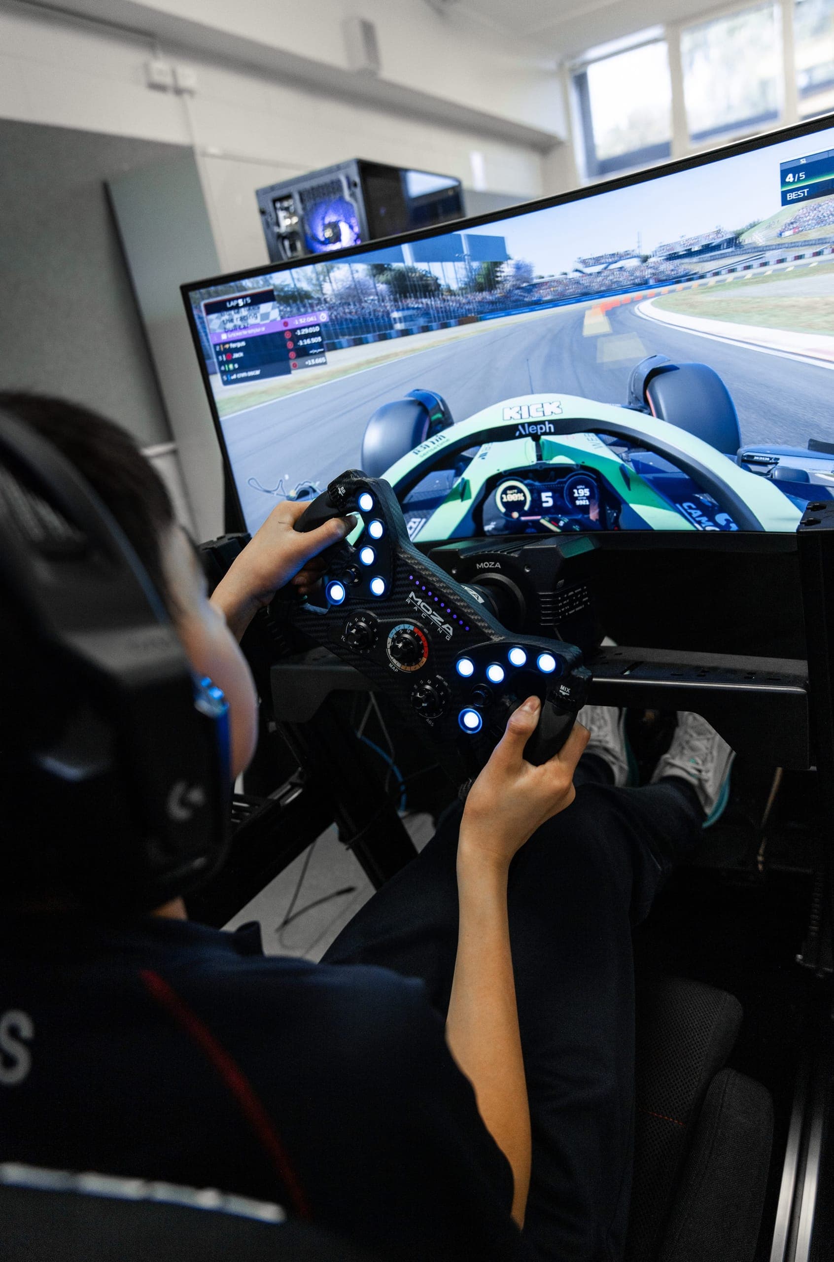 Over a student's shoulder at a simulator wheel, a Formula 1 race on the screen