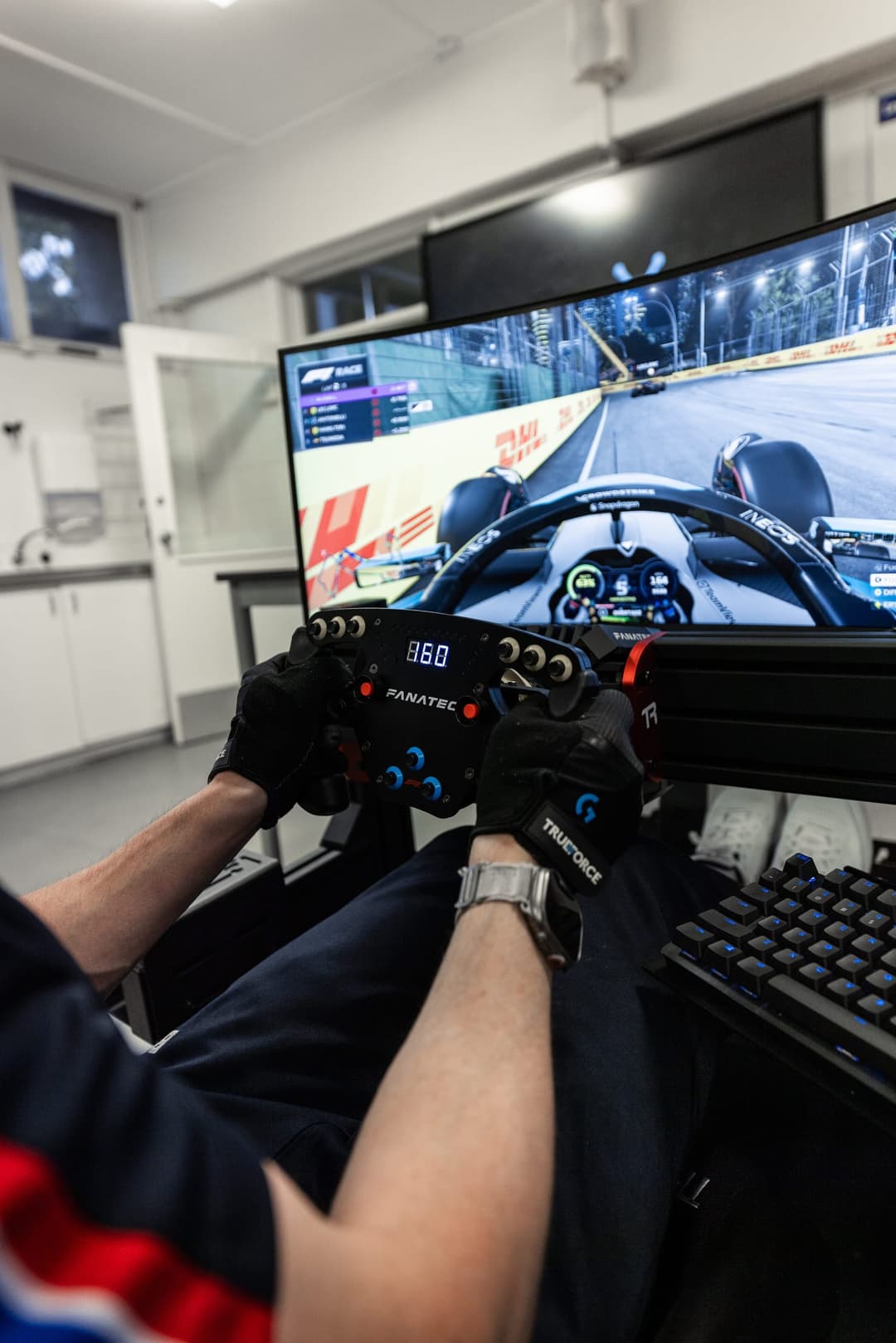 Over the shoulder at a simulator rig, night race on screen and the wheel readout lit