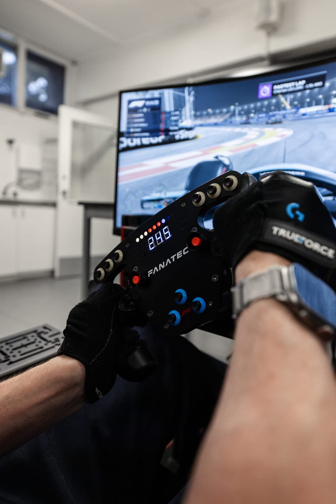Close on a simulator wheel and gloved hands, the race running on the screen behind