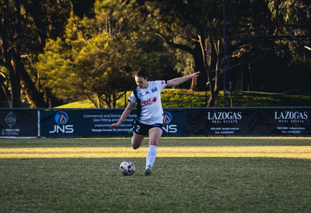 A Calder United player striking a long ball in front of the sponsor boards. Frame 3 of 12. Open it