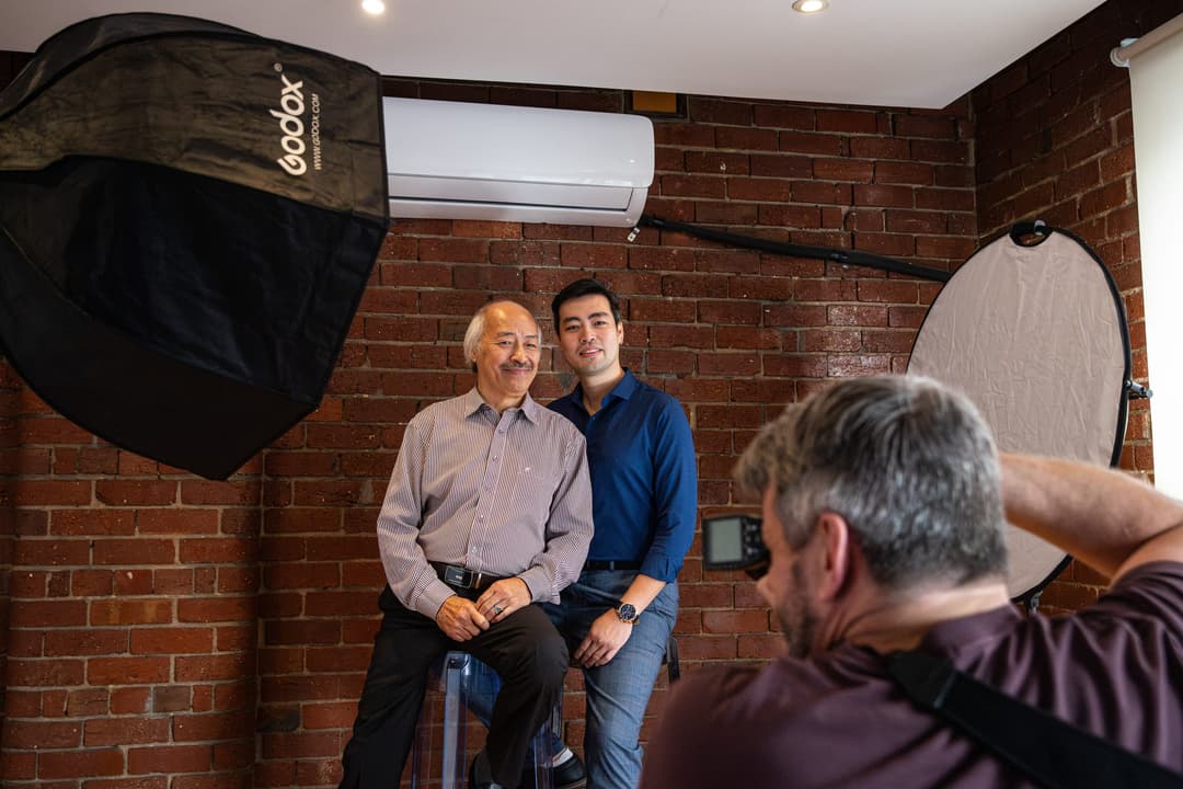Two men posing on stools against a brick wall while the photographer works. Frame 2 of 6. Open it