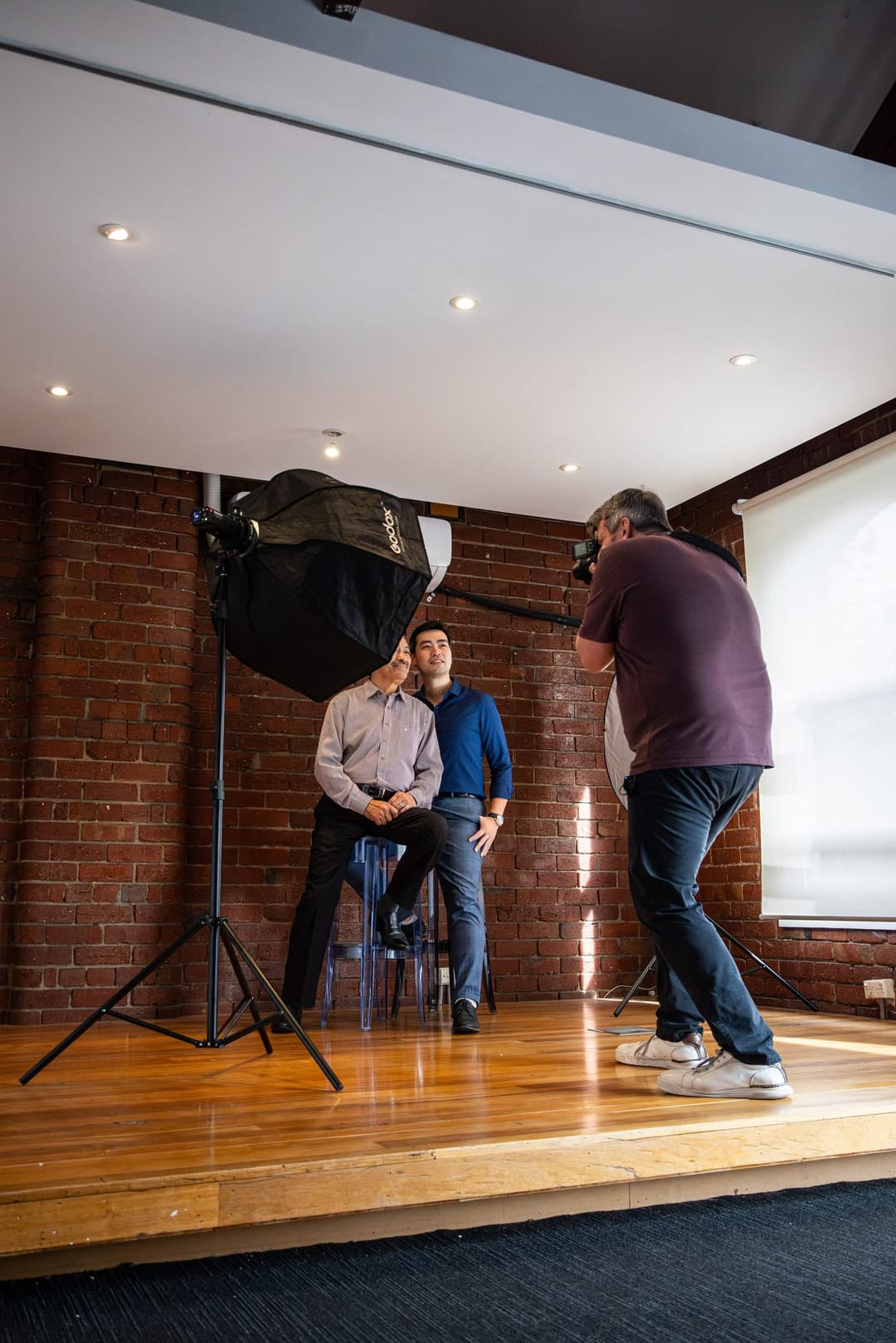 A photographer shooting two men seated on stools against a brick wall