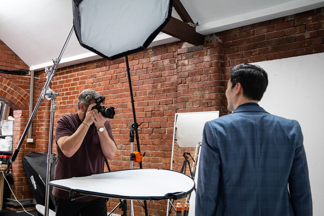 A photographer shooting a man in a blue suit across a reflector in a brick studio