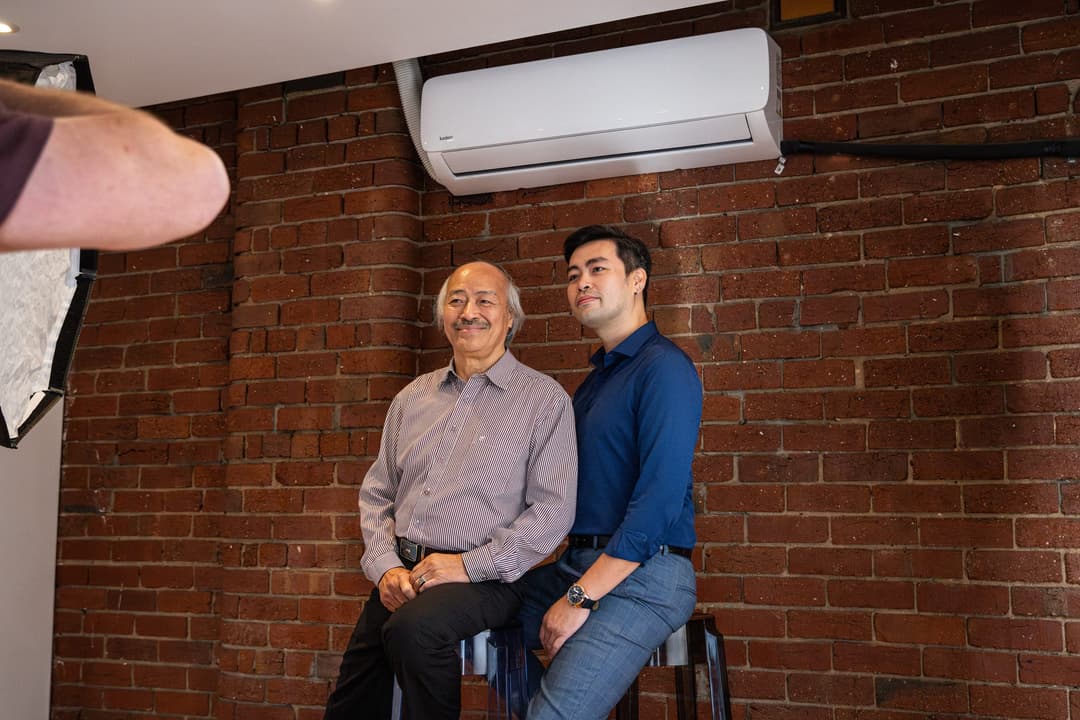 Two men posing on stools against a brick wall, lit by a softbox out of frame